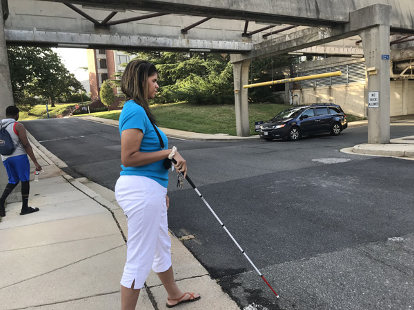 picture shows a student with a white cane standing on the other side of the same crosswalk shown in the previous pictures. To her left, there is a stop sign about 40 feet away, and across the street about 30 feet away is the exit from a parking garage.  A vehicle is waiting to come out of the parking garage.